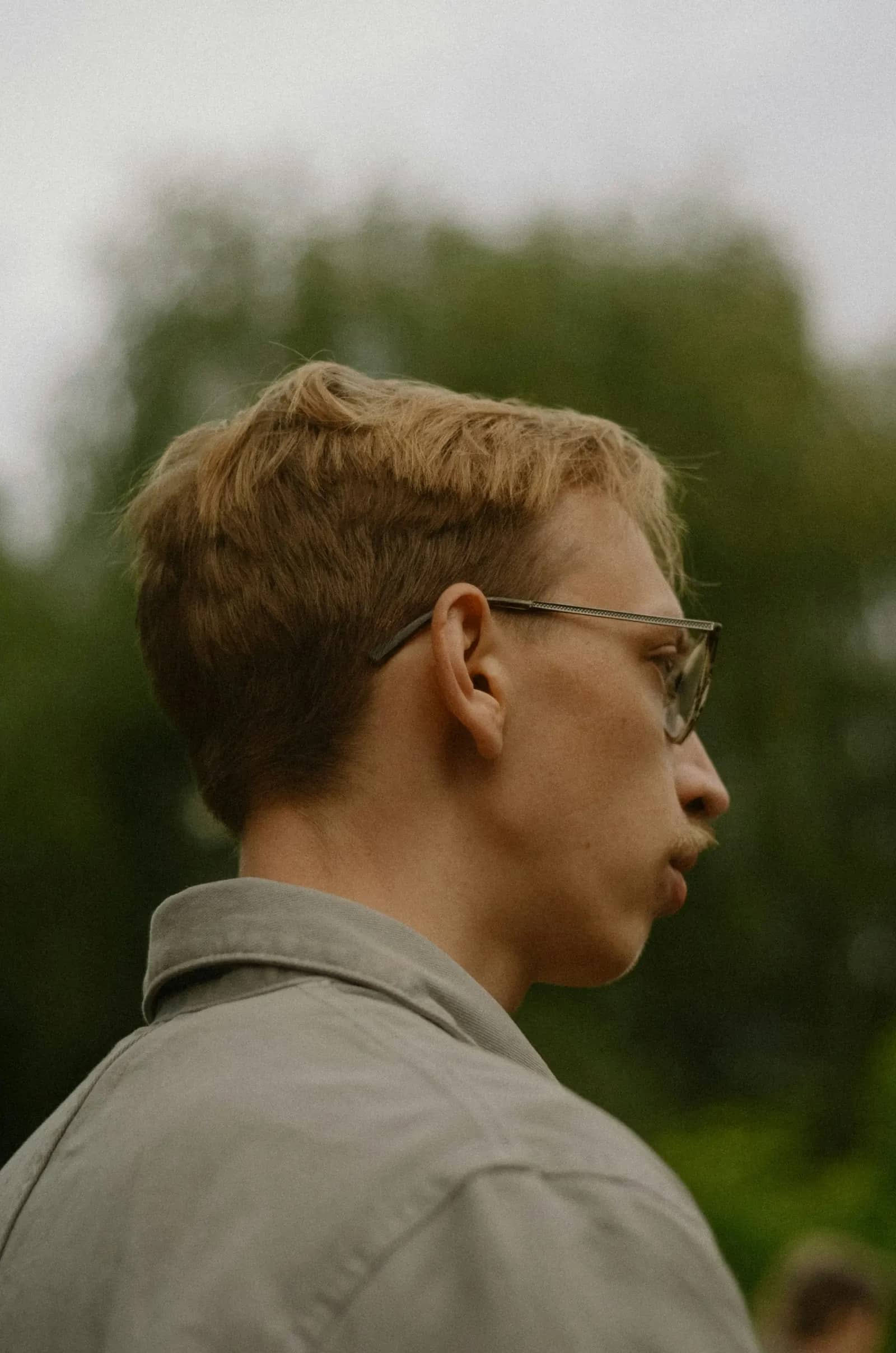 Homme à lunettes, cheveux châtain clair, regardant au loin en extérieur devant un feuillage vert, l'air serein.