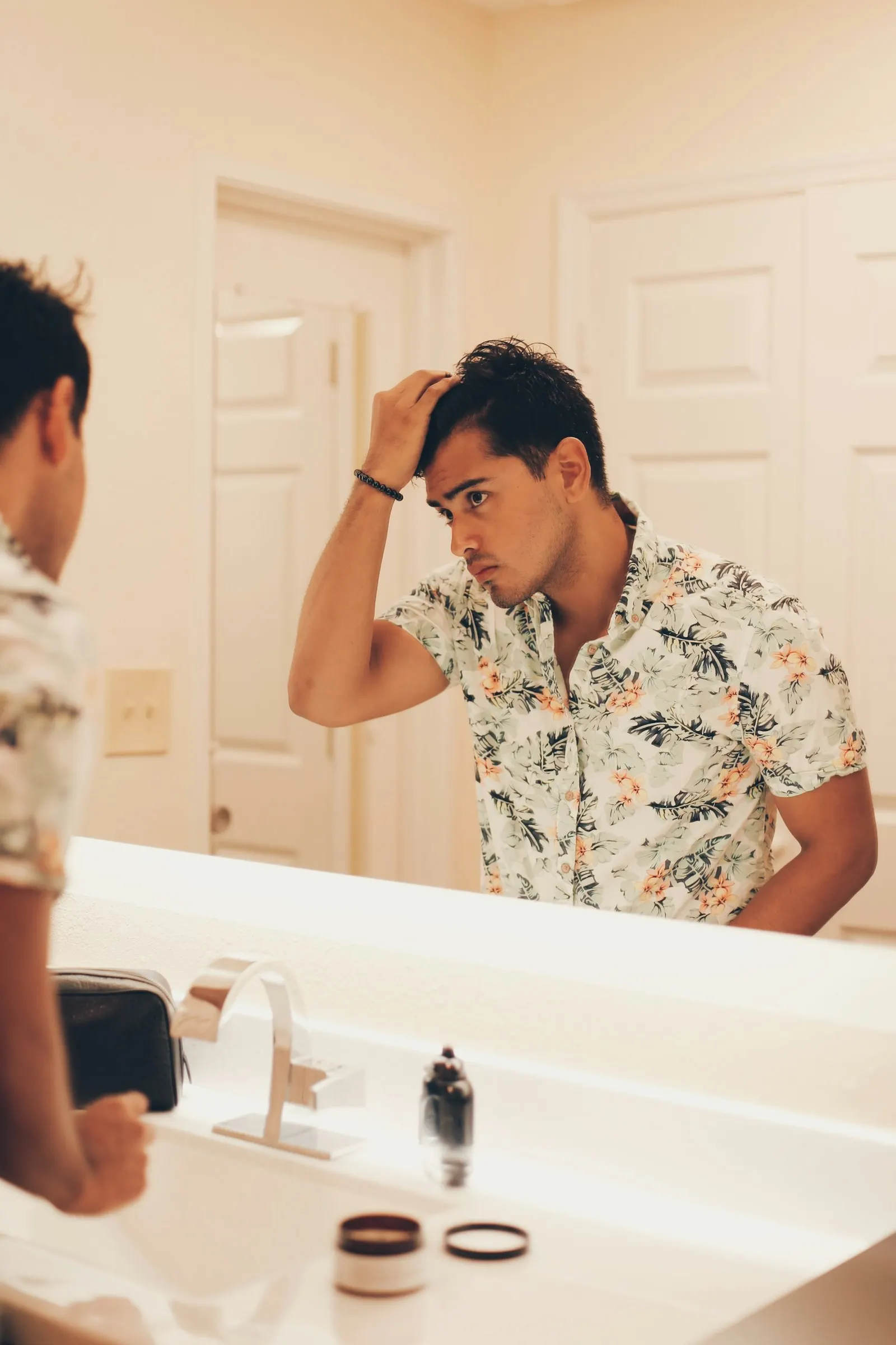 Homme d'une trentaine d'années face au miroir de sa salle de bains, la main dans les cheveux au niveau de la ligne frontale, lumière naturelle douce.
